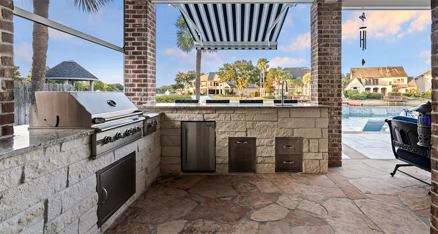 Modern outdoor kitchen with stainless steel grill and storage cabinets, built with stone and brick accents, overlooking a scenic waterfront neighborhood with palm trees and a pool in Frisco, TX.