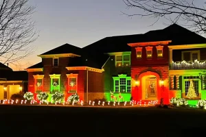 Large two-story home decorated with red and green holiday lighting, illuminated shrubs, wreaths on the windows, and warm lights outlining the entryway and roofline at dusk.