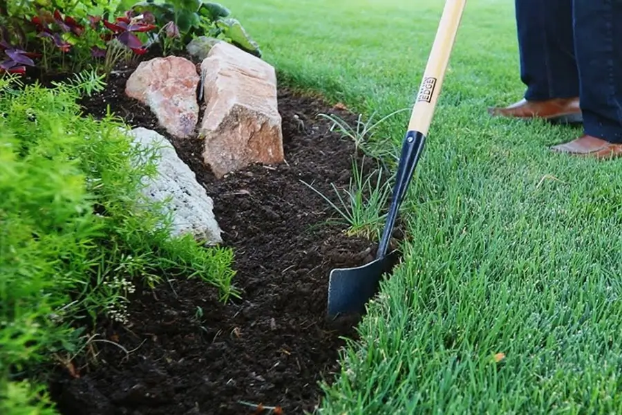 A shovel is creating a clean edge between a green lawn and a freshly prepared planting bed with dark soil, decorative rocks, and small plants.